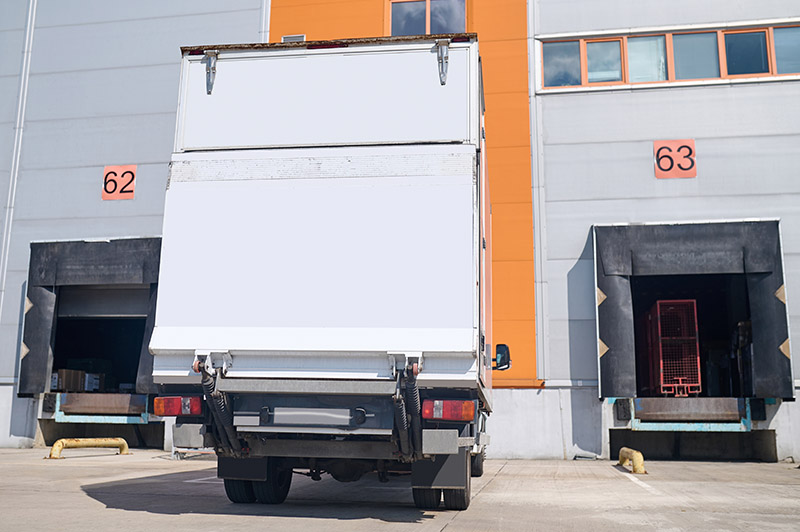 Loading, warehouse. White truck standing outdoors waiting for loading near open porches with numbering in warehouse.
